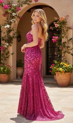 A woman wearing the Blondie Nites 6035BN magenta lace mermaid prom dress with a corset bodice stands outdoors on a stone patio, surrounded by potted plants and arched doorways with blooming flowers.