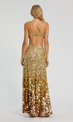 A woman stands facing away, wearing the Long Formal Dress 6077 by Mac Duggal—a gold sequin gown with an open back and a high-waisted, floor-length skirt.