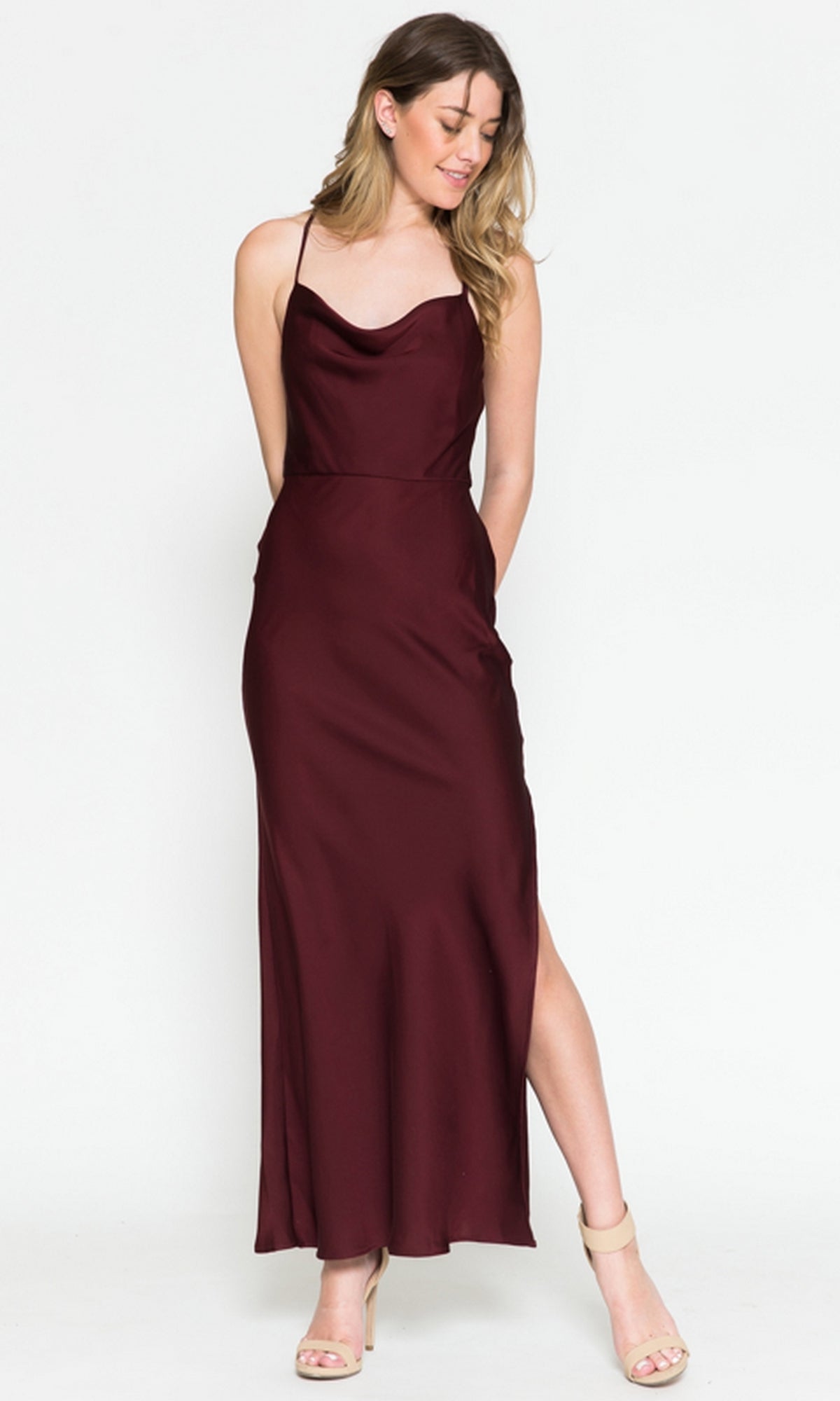 A woman stands against a plain white background wearing the Amelia Couture Cowl-Neck Simple Tea-Length Satin Dress 6115—sleeveless, burgundy satin with spaghetti straps and a side slit.