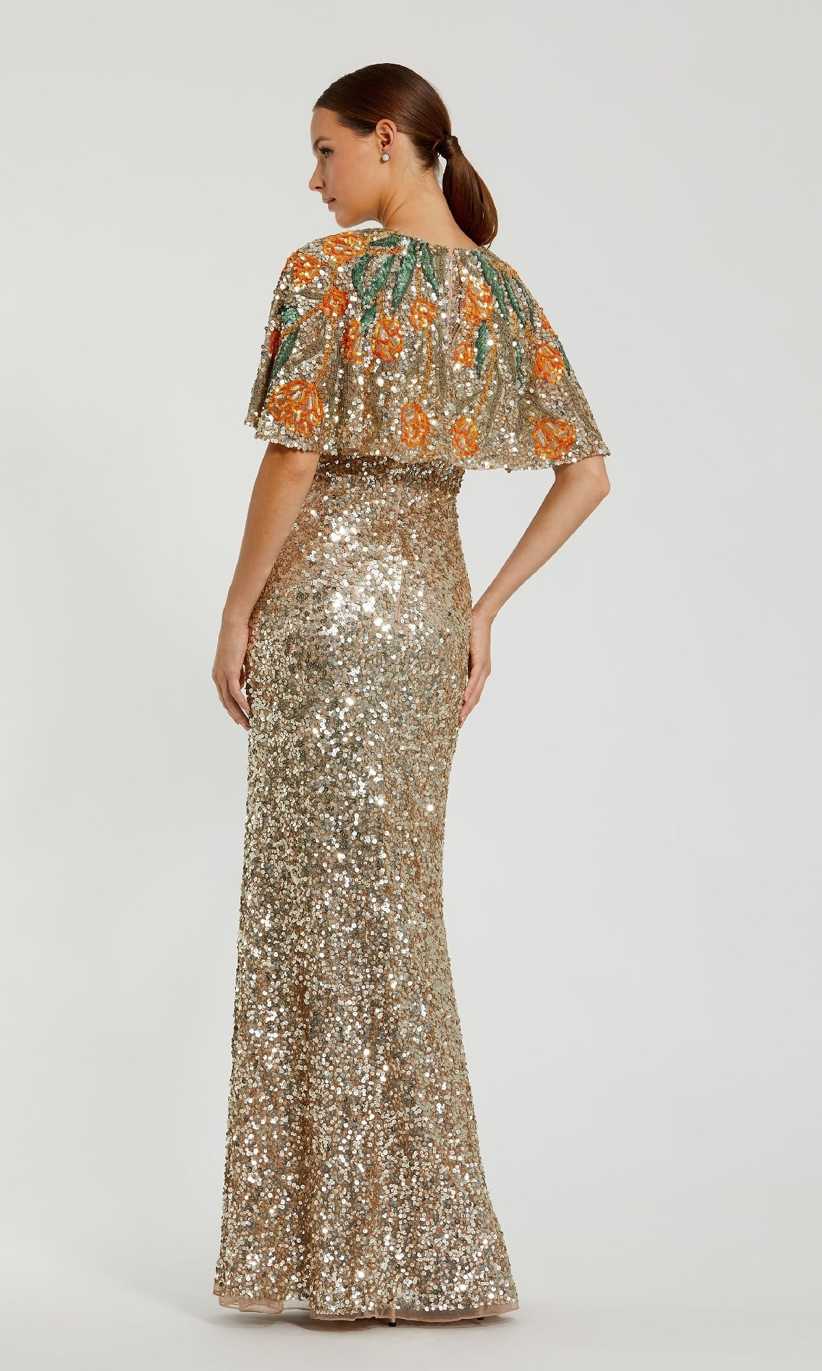A woman stands with her back to the camera in the Caped Gold Sequin Formal Gown: Mac Duggal 6120, featuring a long gold sequin dress and a short embroidered cape with orange floral designs—perfect for mothers of the bride.