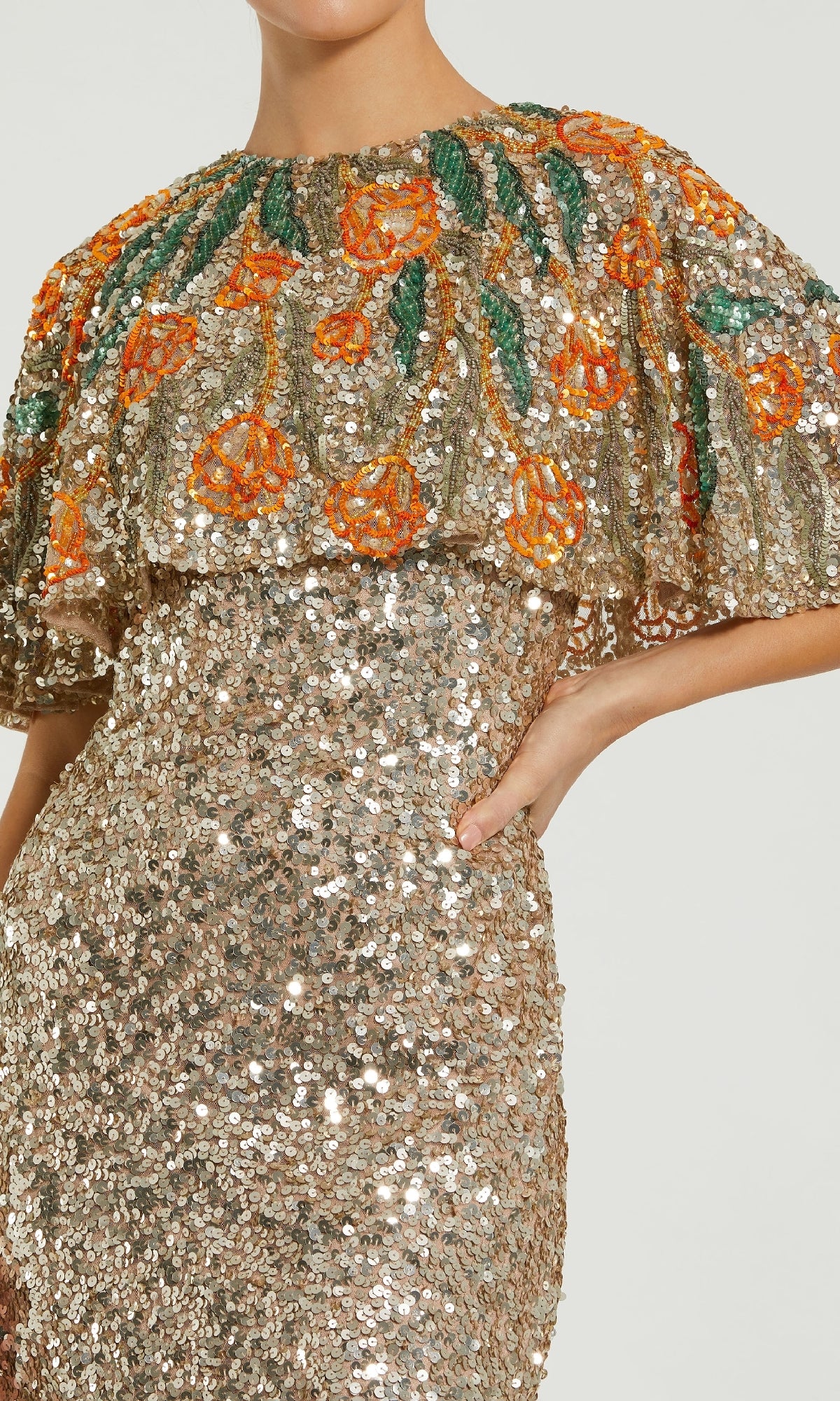 A person wearing the Mac Duggal 6120 Caped Gold Sequin Formal Gown, featuring a shimmering long gold sequin design with a capelet adorned with orange floral embroidery and green leaves—ideal as a mother of the bride dress.