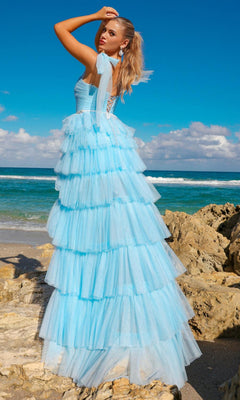 A woman stands on rocks by the ocean in the Bow-Strap Ruffled Long Prom Ball Gown 6125, a light blue tulle dress with ruffles, glancing over her shoulder at the camera beneath a partly cloudy sky.