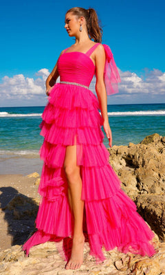 A woman stands barefoot on a rocky beach in the Bow-Strap Ruffled Long Prom Ball Gown 6125, featuring bright pink tiers, an asymmetrical hem, and a belted waist. The ocean and sky provide a stunning backdrop.