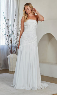 A woman stands indoors on a light rug, wearing the Amelia Couture 6132 long prom dress with a strapless neckline and flattering ruching, framed by a large vase of branches and a white arched alcove in the background.