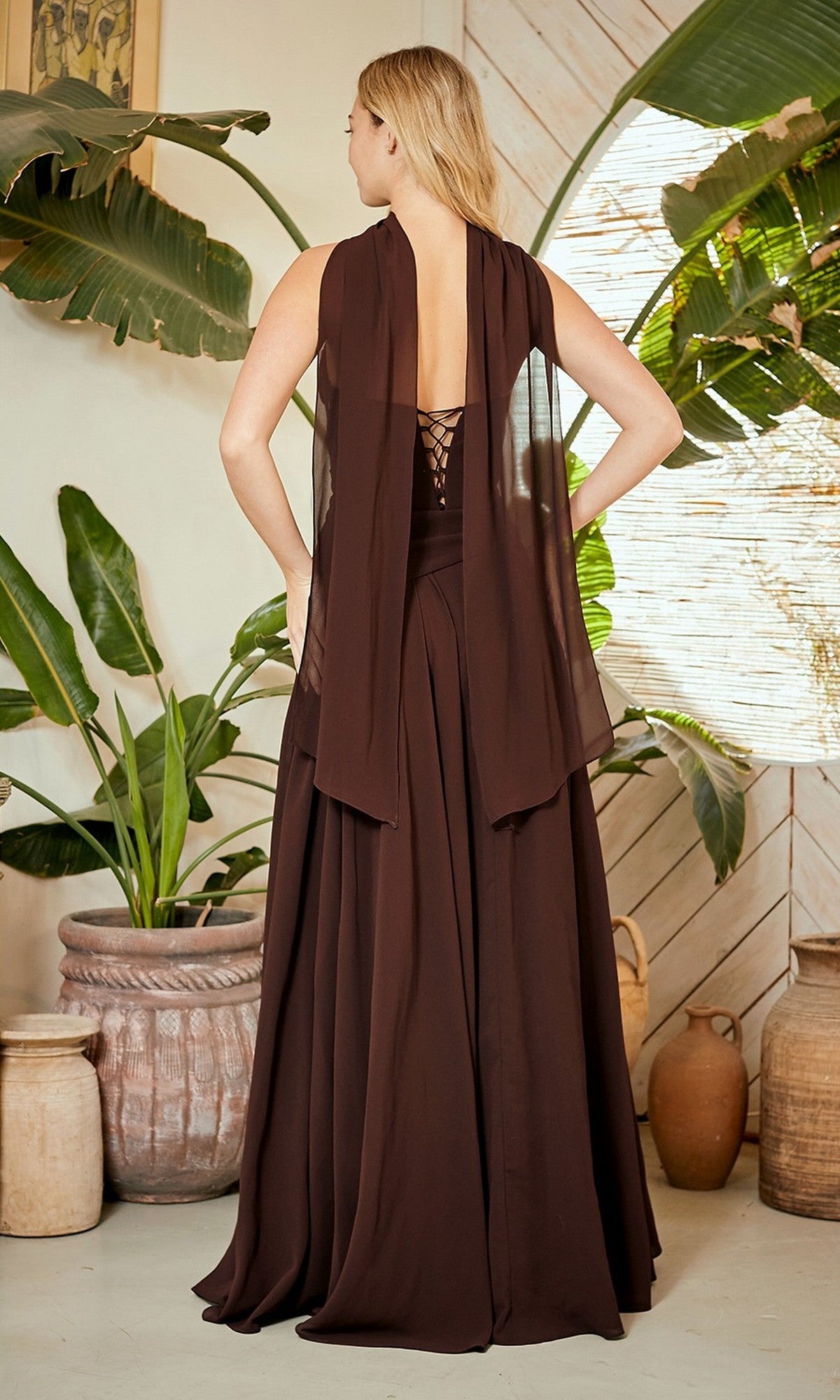 A woman stands indoors in the Amelia Couture 6133 long prom dress, featuring a corset back and sheer panels, surrounded by large potted plants and rustic pottery.