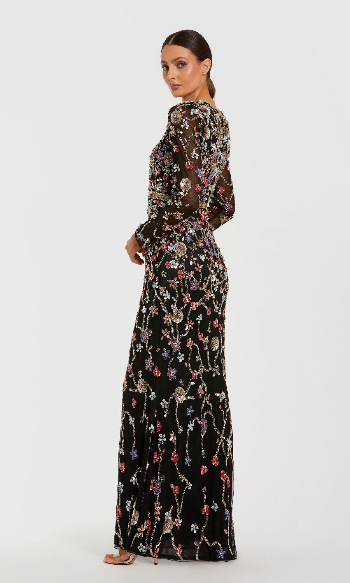 A woman stands in a side pose against a white background wearing the Mac Duggal 6135 Long Black Floral Formal Dress with colorful embroidery, beading, sheer long sleeves, and open-toe heels.