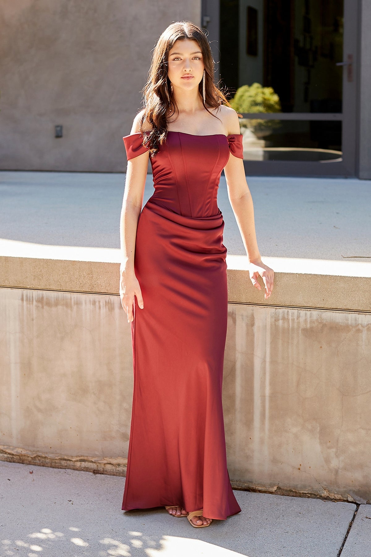 A woman wearing a burgundy, off-the-shoulder Amelia Couture 6151 gown with a corset-style bodice and a floor-length skirt.