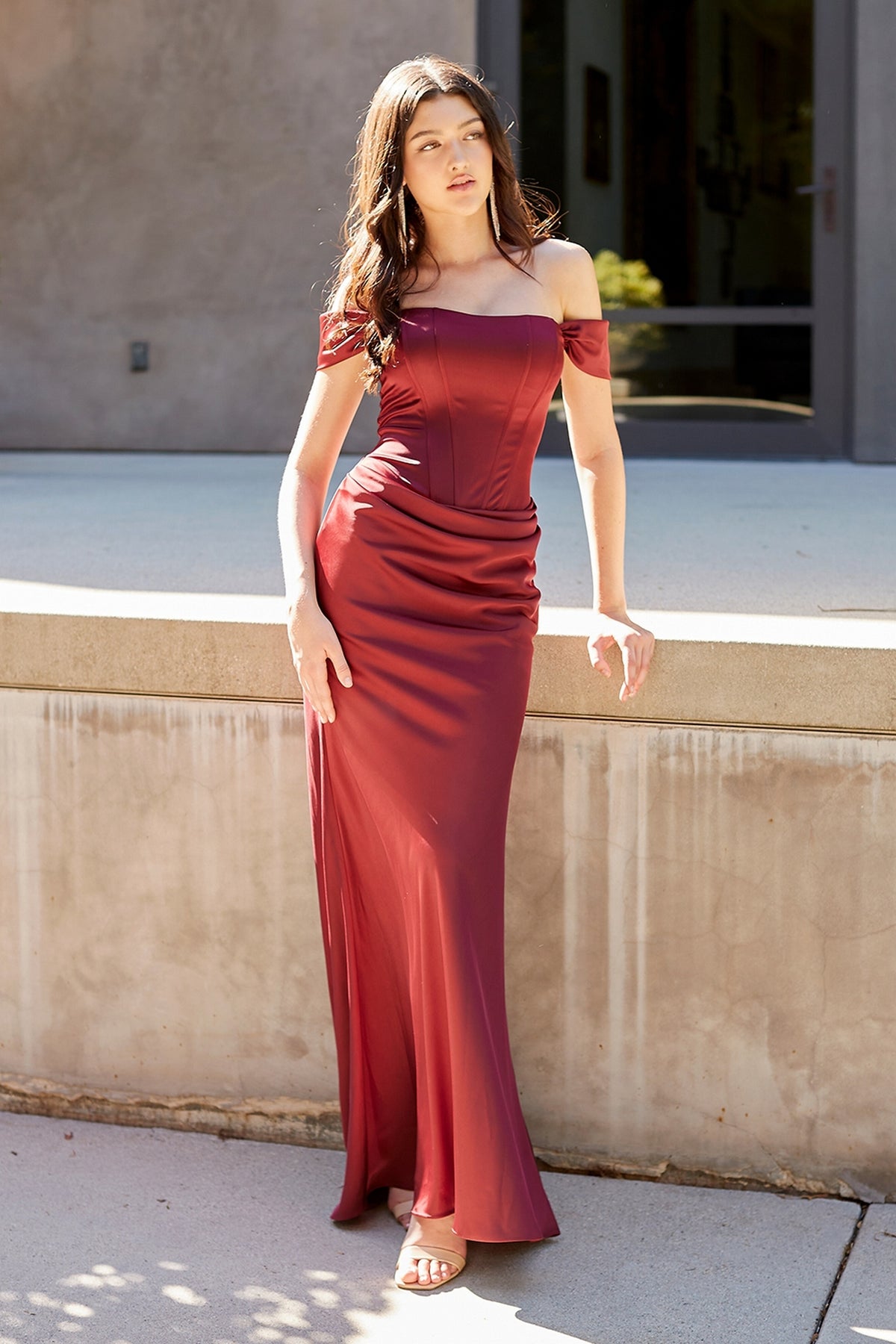 A woman wearing a burgundy off-the-shoulder Amelia Couture 6151 floor-length prom dress with a corset-style bodice.
