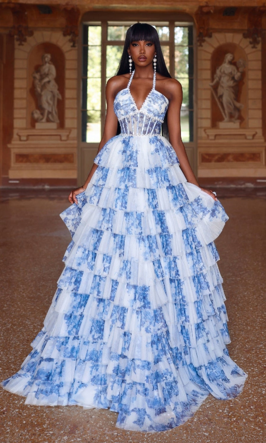A woman stands indoors wearing the Alyce 61730 Ruffled Long Print Prom Dress, featuring a white and blue print, halter neckline, and structured bodice. She accessorizes with long beaded earrings; statues and large windows are visible in the background.