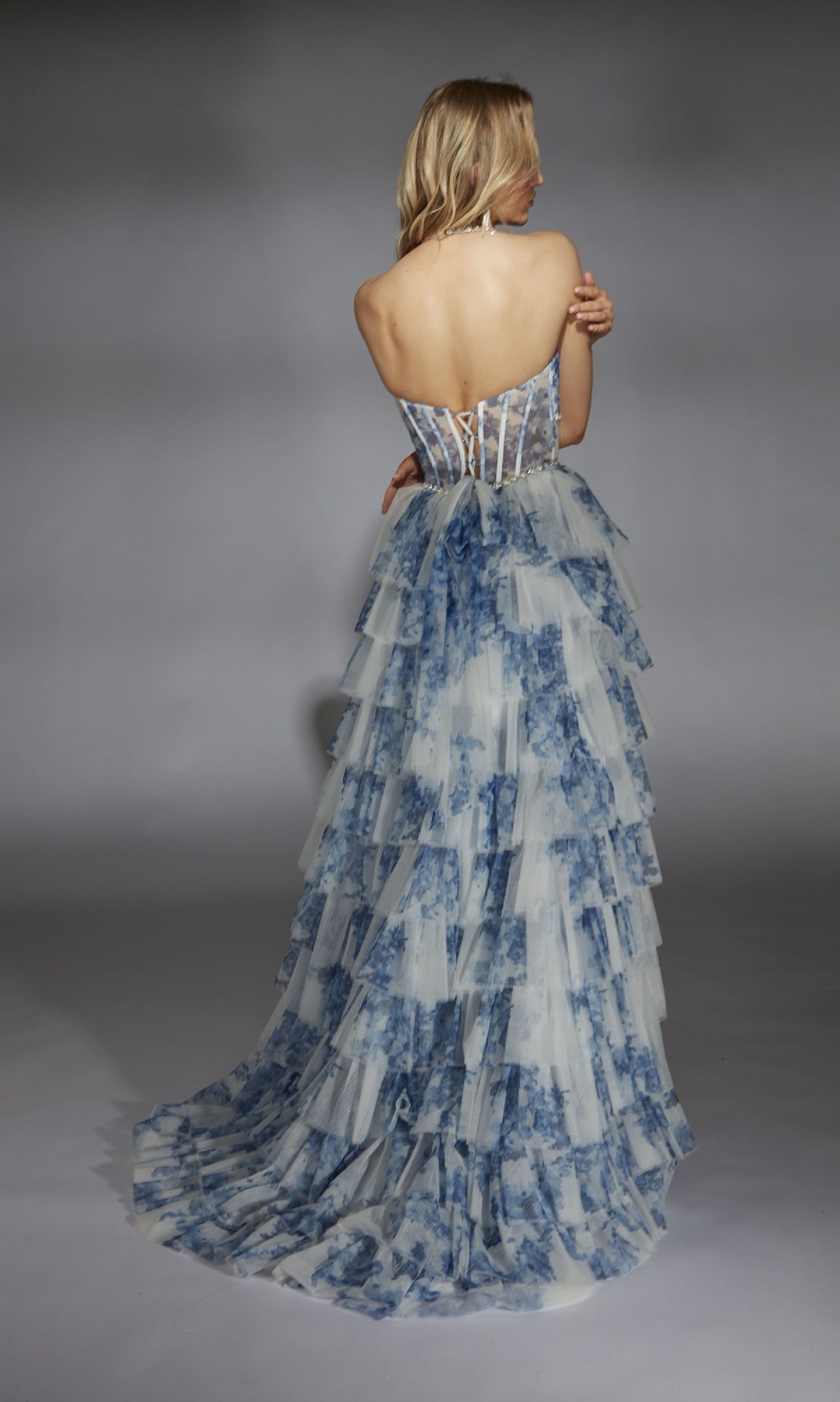 A woman faces away, wearing the Alyce 61730 Ruffled Long Print Prom Dress, featuring blue and white tiered ruffles and a sheer corset back, set against a plain studio backdrop.