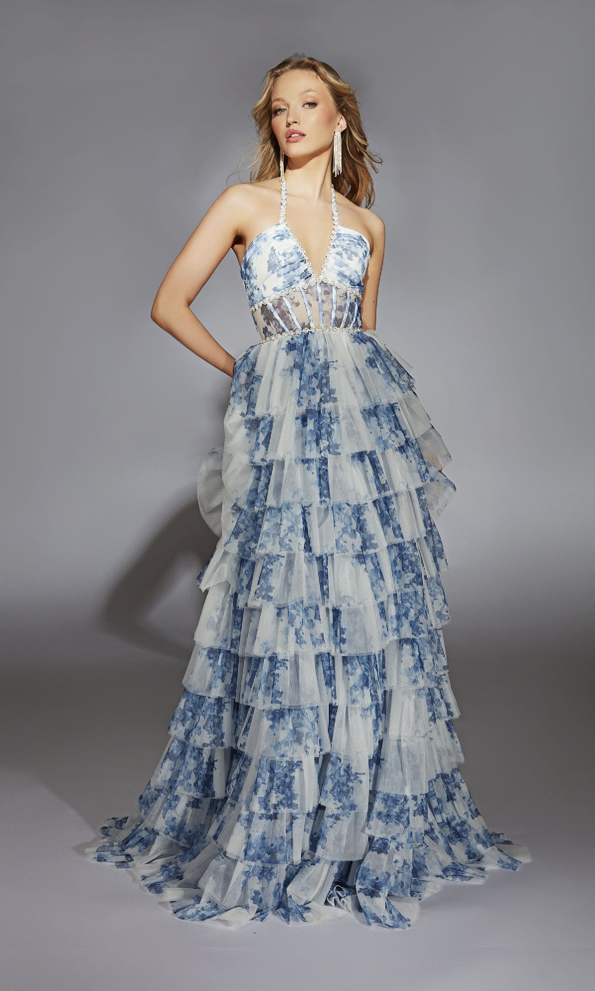 A woman wearing the Alyce 61730 Ruffled Long Print Prom Dress, featuring a sleeveless halter-neck, sheer bodice, tiered ruffled skirt, and blue floral pattern stands against a gray background.