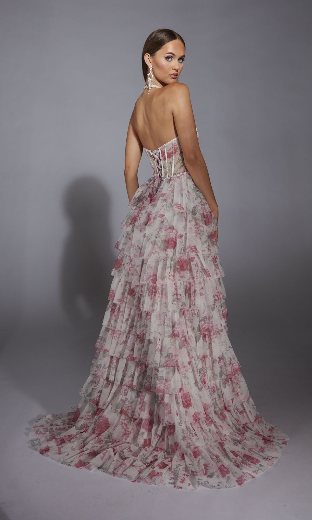 A woman wearing the Alyce 61730 Ruffled Long Print Prom Dress—strapless, floor-length, and tiered with a pink floral pattern—stands turned slightly to the side against a plain gray background.