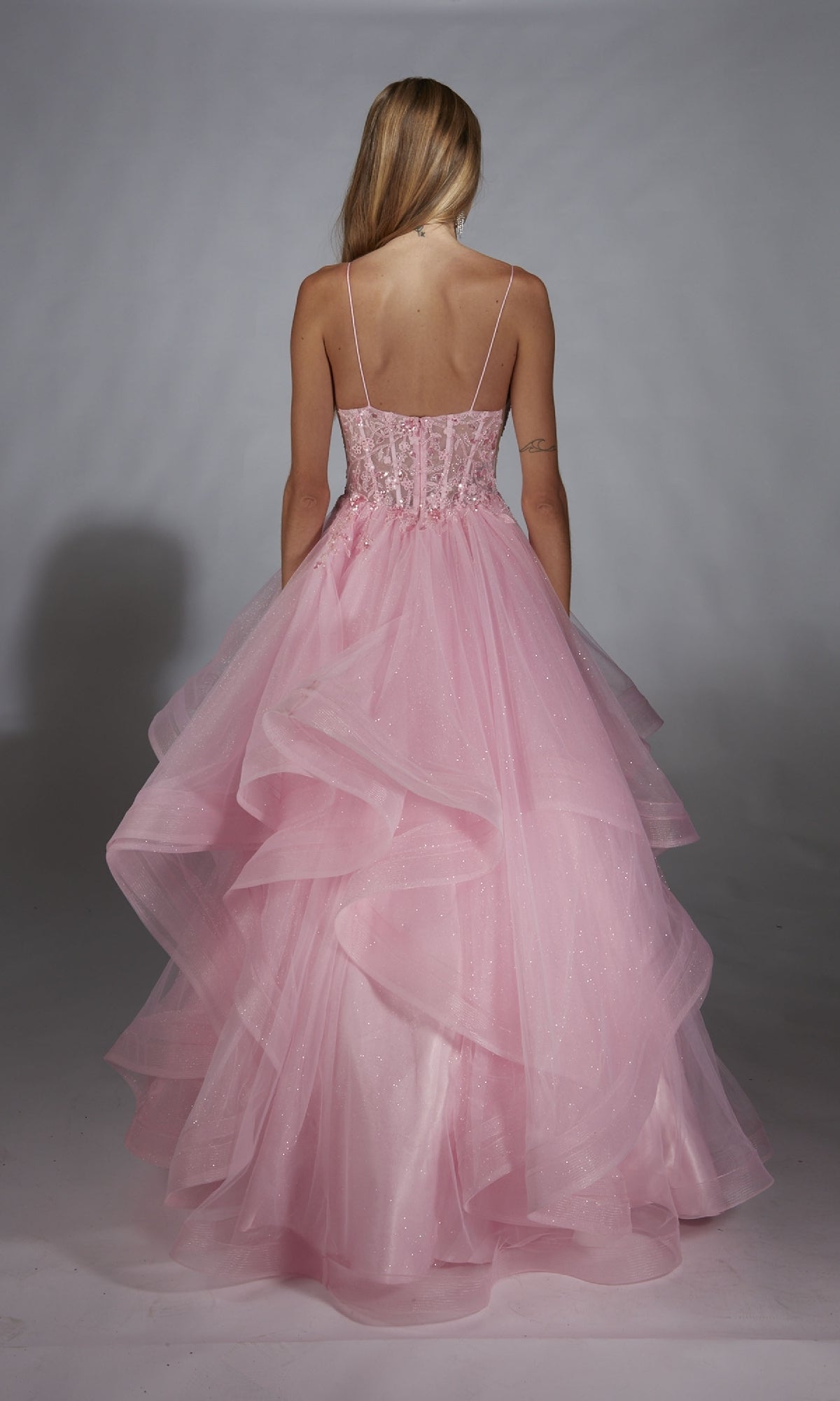 A woman in the Alyce 61731 Long Prom Dress, featuring a lace embroidered corset and thin straps, faces away from the camera as her light pink layered tulle gown flows elegantly against a plain backdrop.