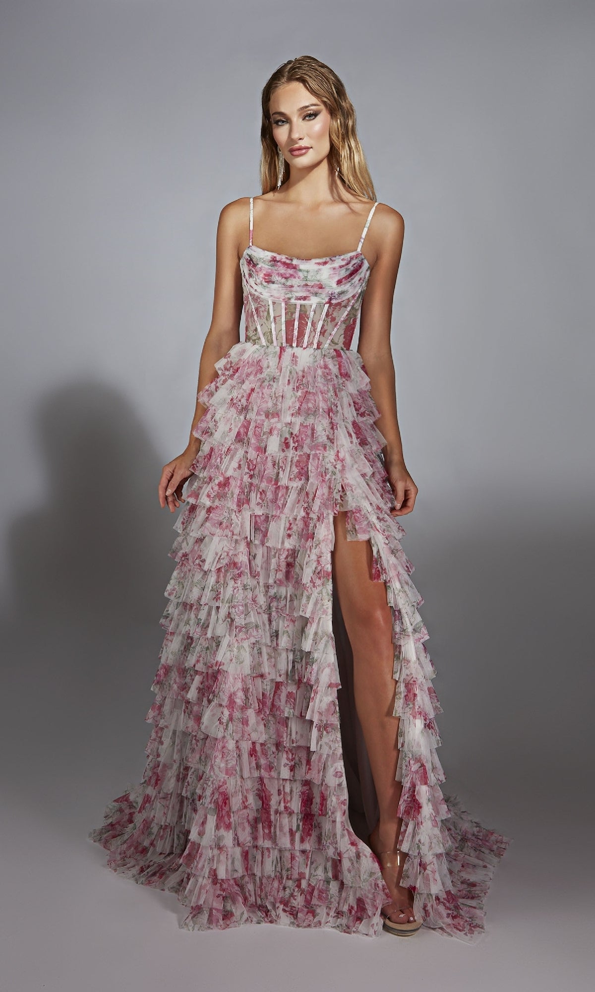 A woman models the Alyce 61732, a sleeveless, floor-length pink and white prom dress with floral patterns, ruffles, a corset bodice, and high slit, standing against a plain backdrop.