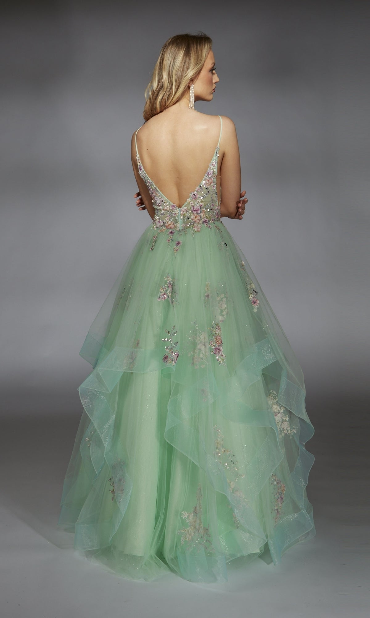 A woman showcases the Alyce 61735 long prom dress, featuring a light green layered tulle skirt, floral embellishments, thin straps, and a deep V-back, as she stands facing away against a neutral background.
