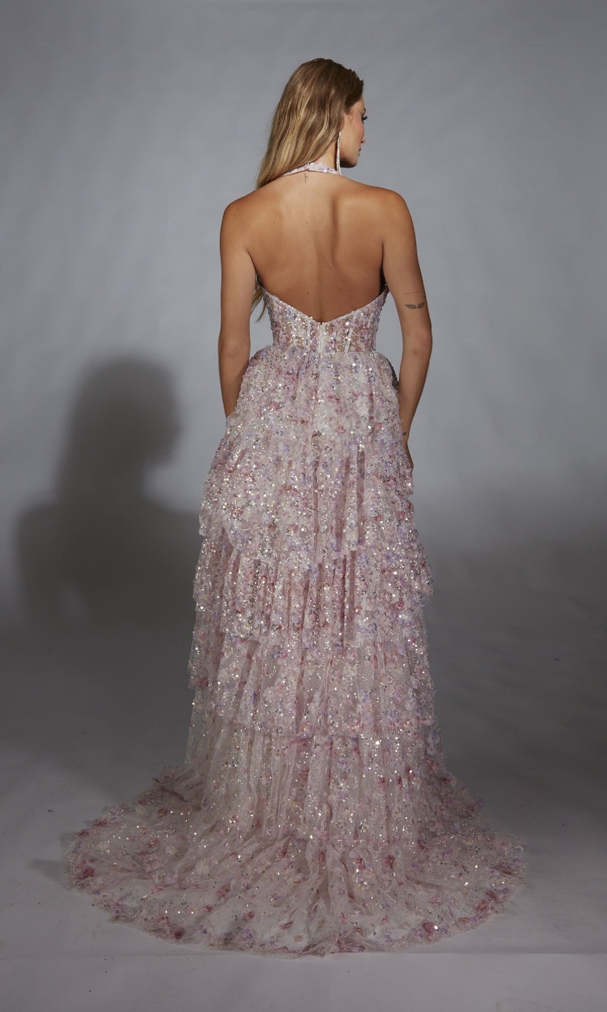 A woman faces away, wearing the Alyce 61740 long prom dress with layered ruffles and sequins in light pink and white, set against a plain gray background.