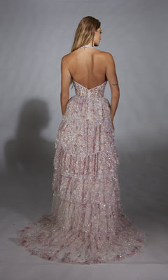 A woman faces away, wearing the Alyce 61740 long prom dress with layered ruffles and sequins in light pink and white, set against a plain gray background.