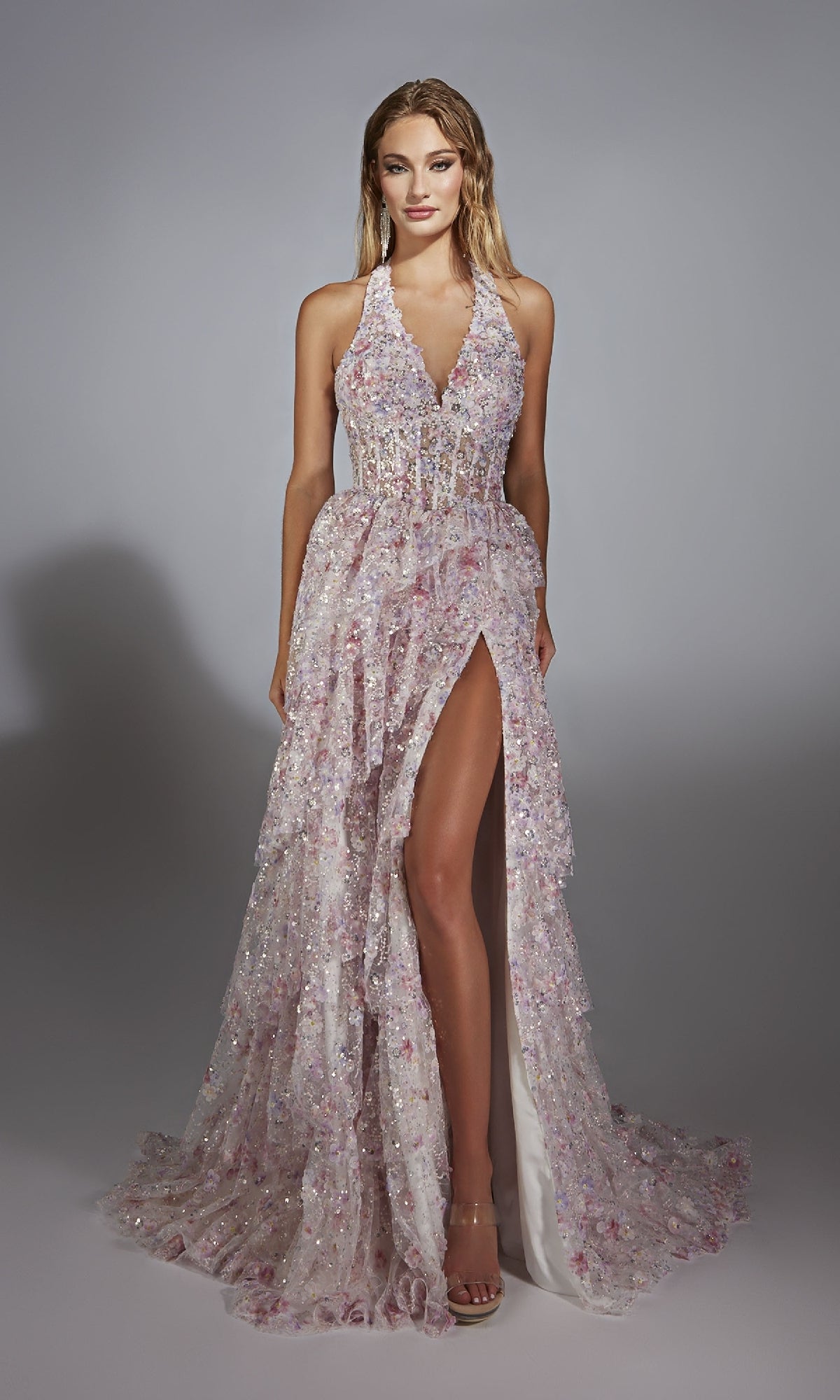A woman poses in a studio wearing the Alyce 61740 long prom dress—a sleeveless, floor-length gown with a high slit and sheer sparkly floral design in pale pink and lavender.