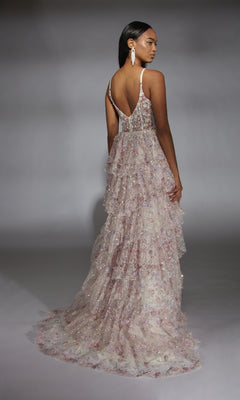 A woman faces away, wearing the Alyce 61745 long prom dress—a tiered, floor-length glitter tulle gown with floral embellishments, thin straps, and a corset bodice—set against a plain gray background.