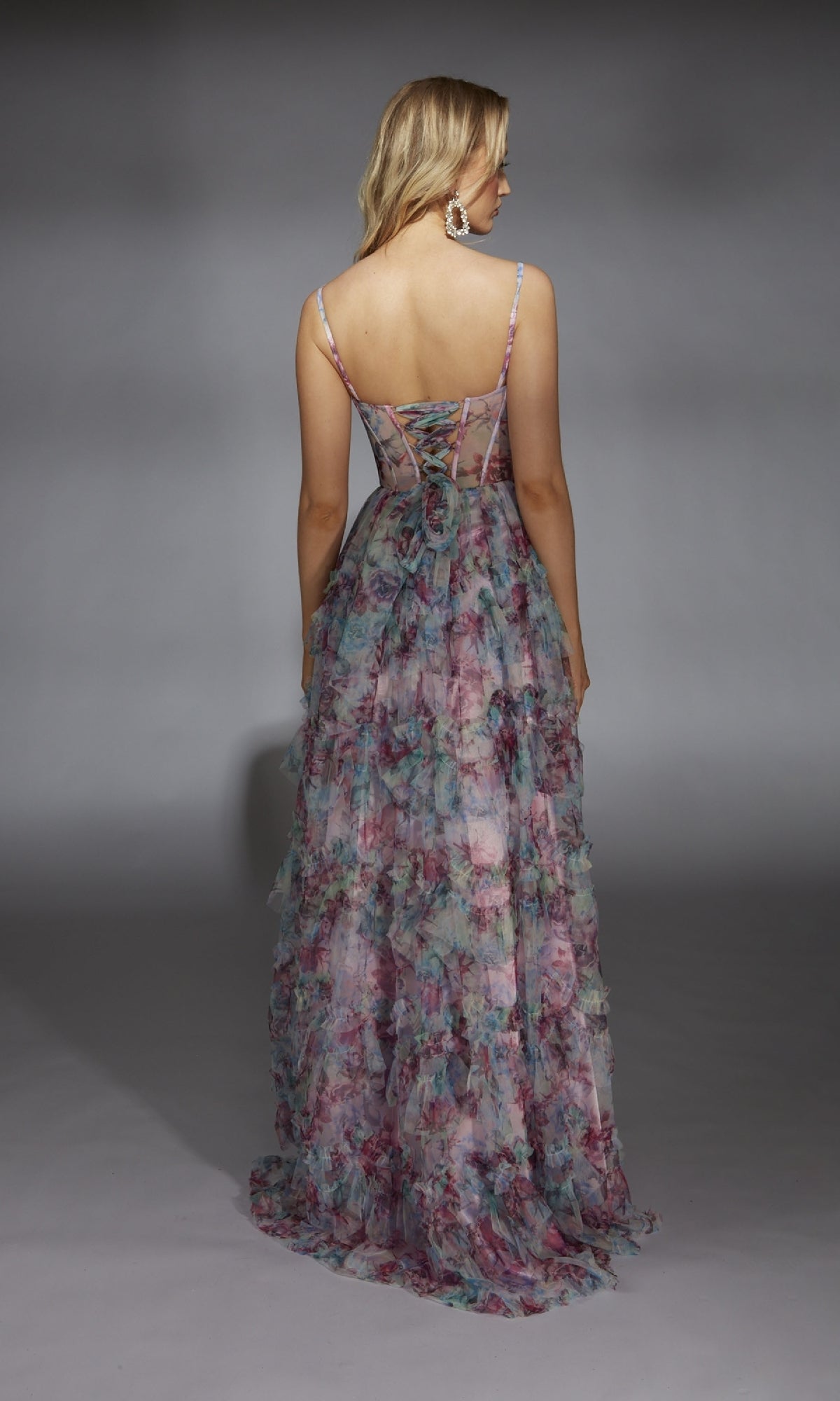 A woman faces away, wearing the Alyce 61748 long prom dress with a high-leg slit, lace-up back detail, and floral patterns, set against a plain gray background.