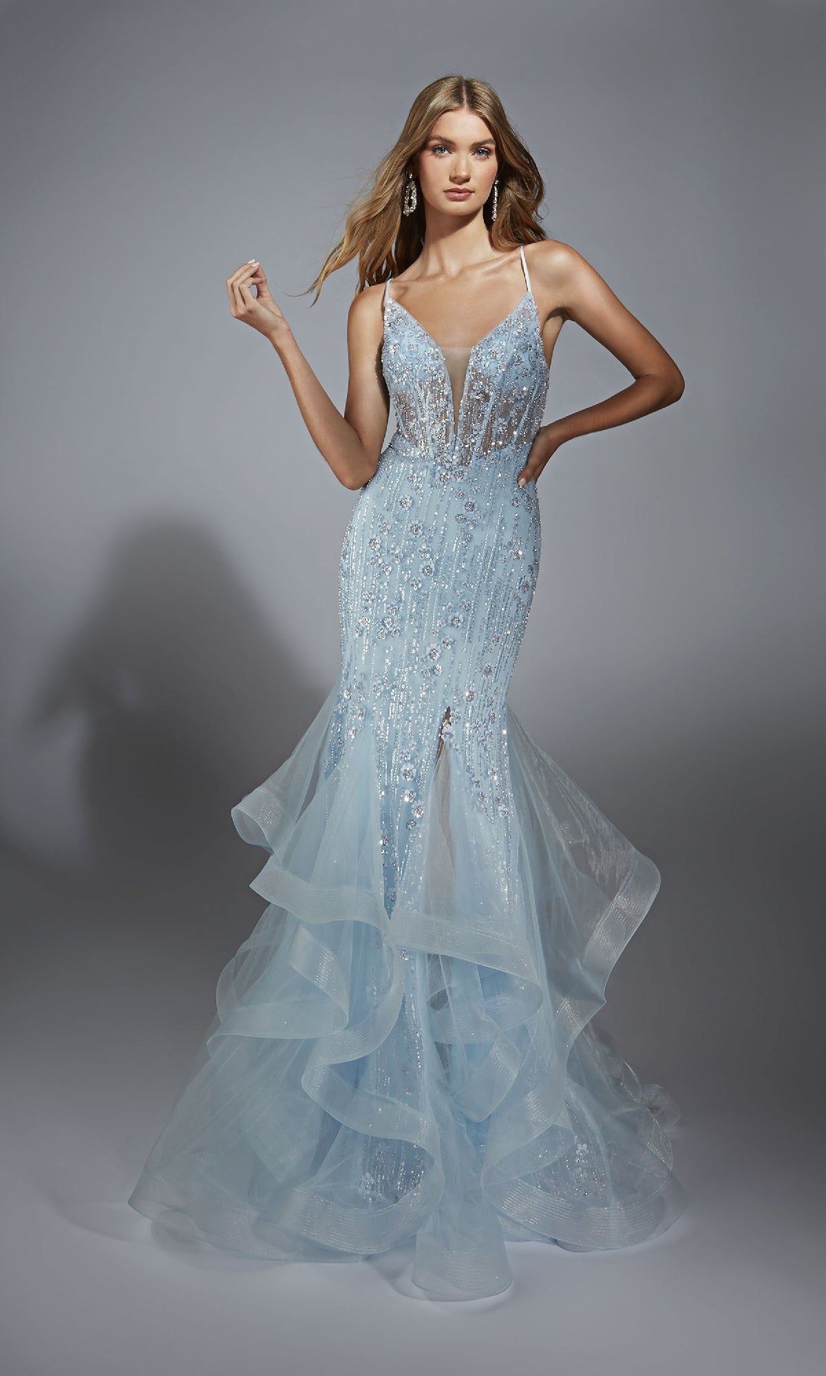 A woman stands against a gray background in the Alyce 61749 long prom dress, featuring a light blue beaded mermaid silhouette with a lace-up corset bodice, deep V-neckline, and layered tulle skirt.