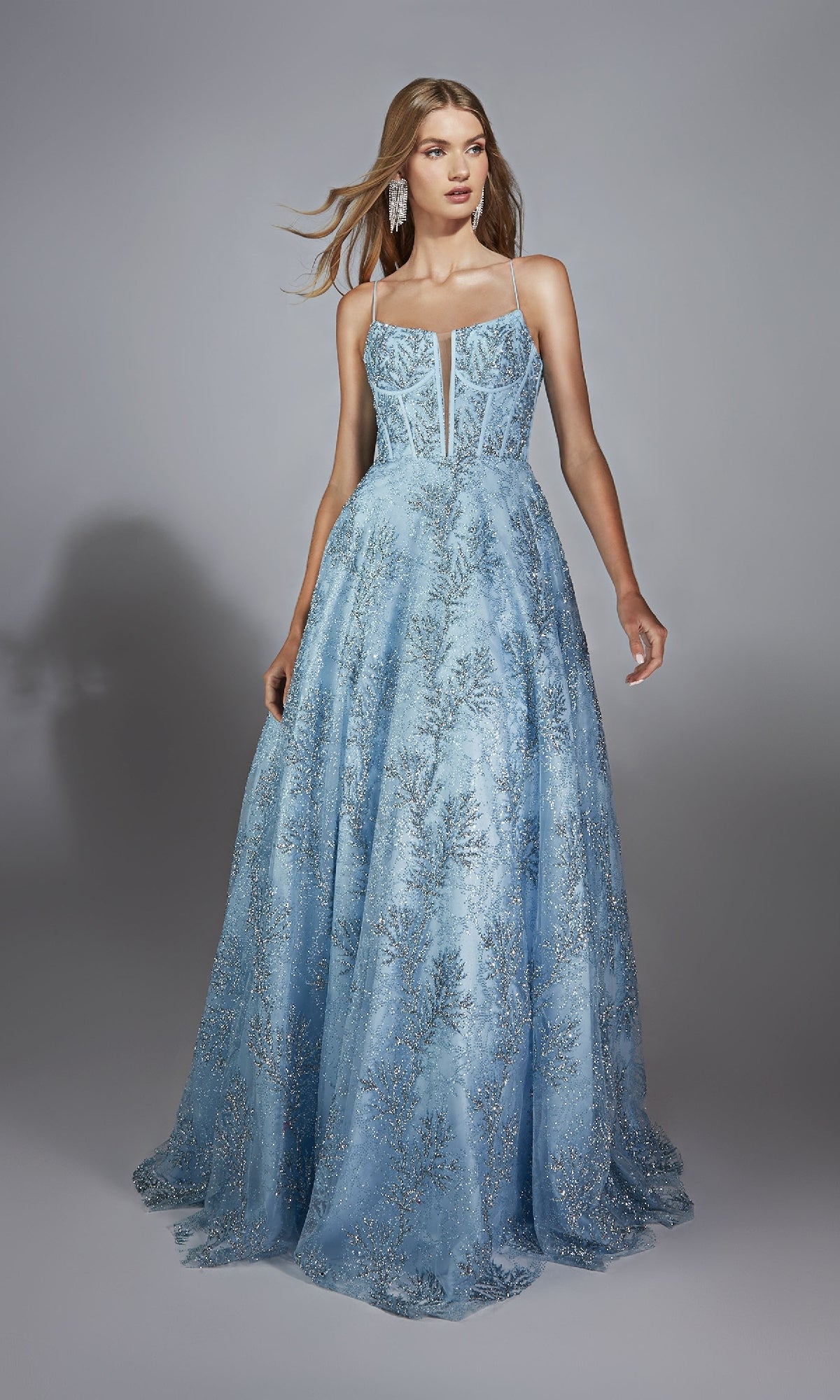 A woman models the Alyce 61751 long prom dress, featuring a light blue color, corset bodice, thin straps, glitter embellishments, and a floor-length design against a plain gray background.