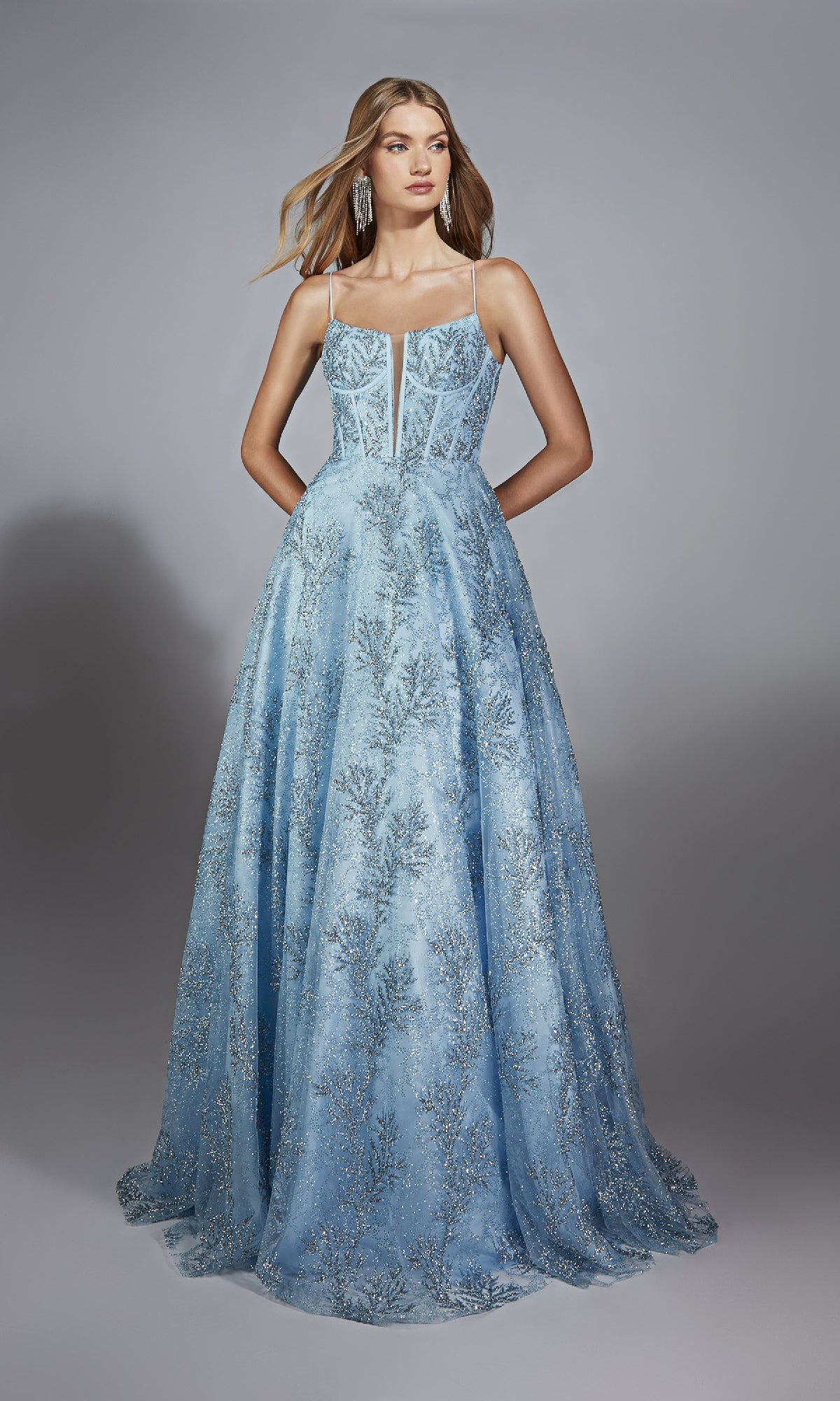 A woman models the Alyce 61751, a floor-length light blue prom dress with silver embroidery, thin straps, and a corset bodice, posing elegantly against a plain gray background.
