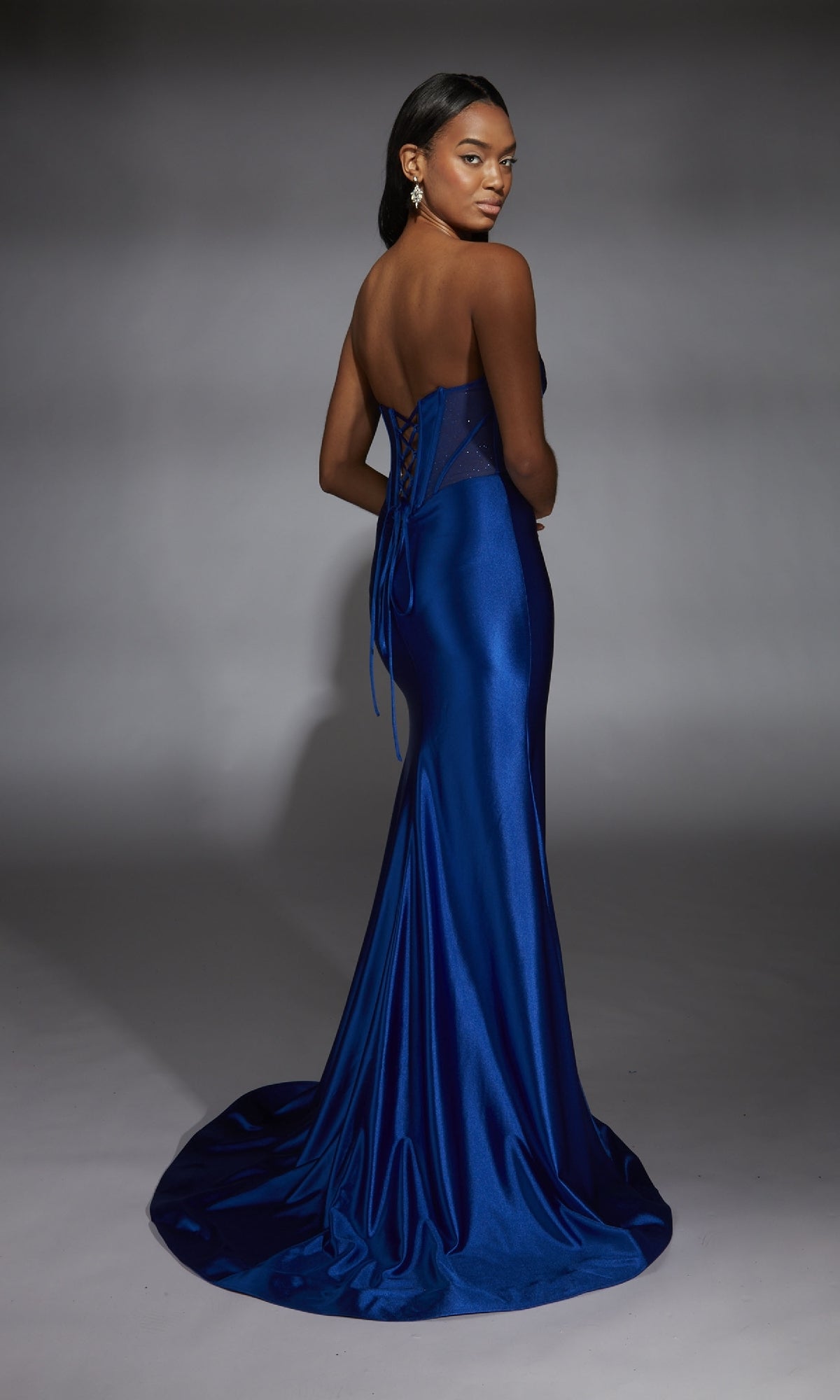 A woman poses in the Alyce 61754 long prom dress—a fitted, strapless royal blue satin gown with a lace-up back—looking over her shoulder against a gray studio background.