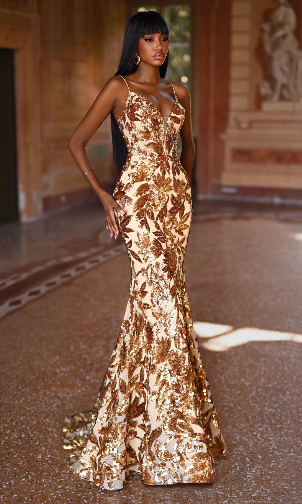 A woman in the Alyce 61755 Lace-Up Long Sequin Prom Dress, featuring fitted gold and bronze leaf patterns, stands indoors on a patterned floor with ornate walls behind her.