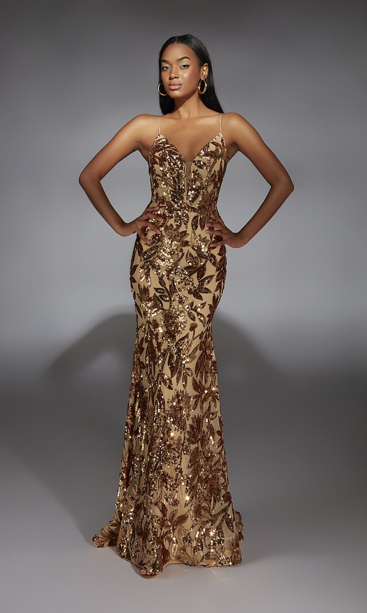 A woman stands with hands on hips in the Lace-Up Long Sequin Prom Dress: Alyce 61755, a fitted, floor-length gold gown with sequined floral patterns, against a plain gray background.