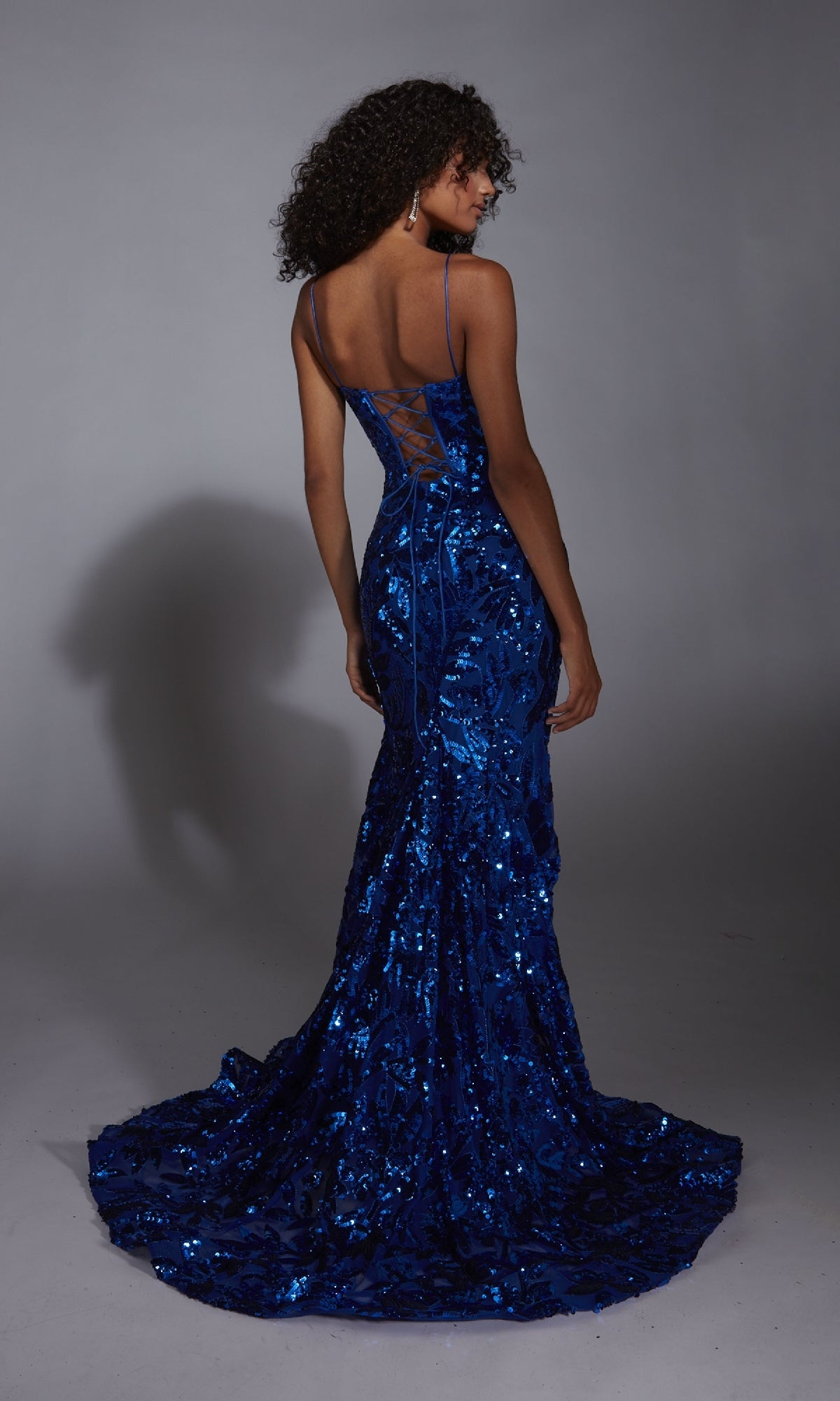 A woman in the Lace-Up Long Sequin Prom Dress: Alyce 61755, featuring a floor-length blue sequin design, fitted bodice, and lace-up back, stands facing away from the camera.