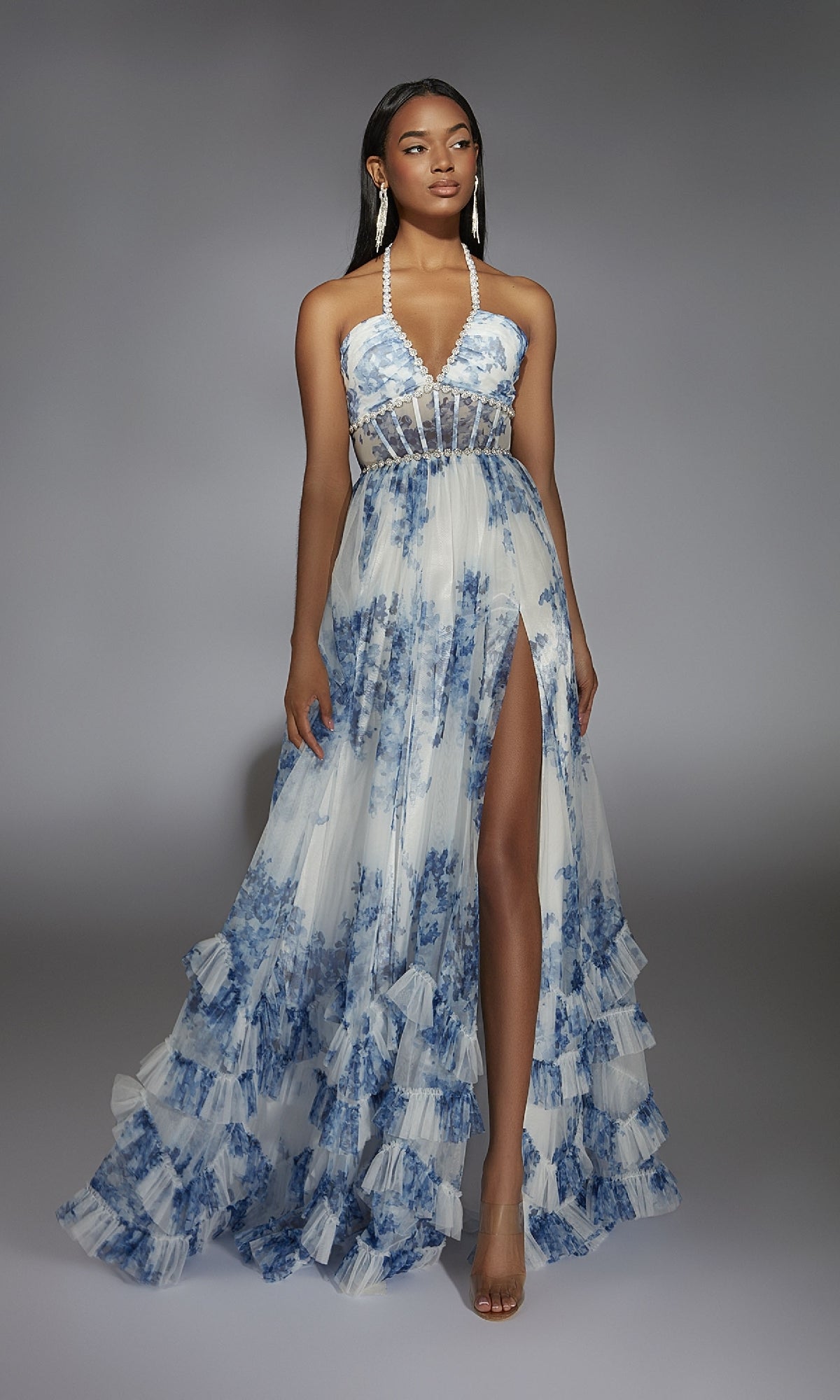 A woman in the Alyce 61757 long prom dress, a blue and white floral halter gown with a sheer bodice, high slit, ruffled hem, and clear heels stands against a gray background.