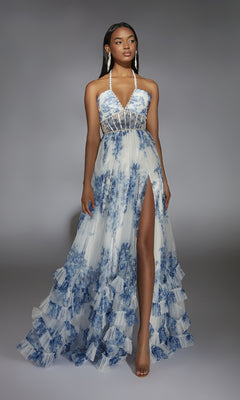 A woman in the Alyce 61757 long prom dress, a blue and white floral halter gown with a sheer bodice, high slit, ruffled hem, and clear heels stands against a gray background.
