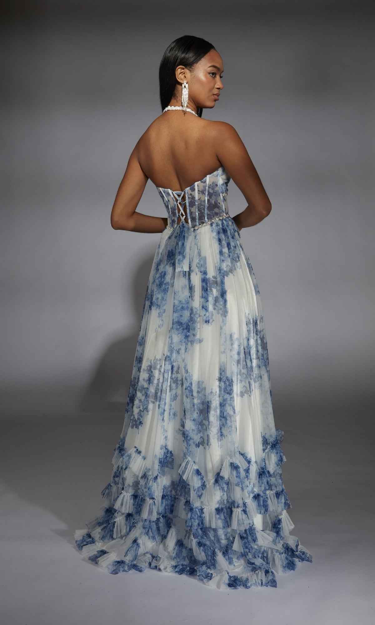 A woman stands against a gray background in the Alyce 61757 long prom dress—a strapless, floor-length white gown with blue floral prints and a lace-up back, offering a modern take on classic 80’s prom style.