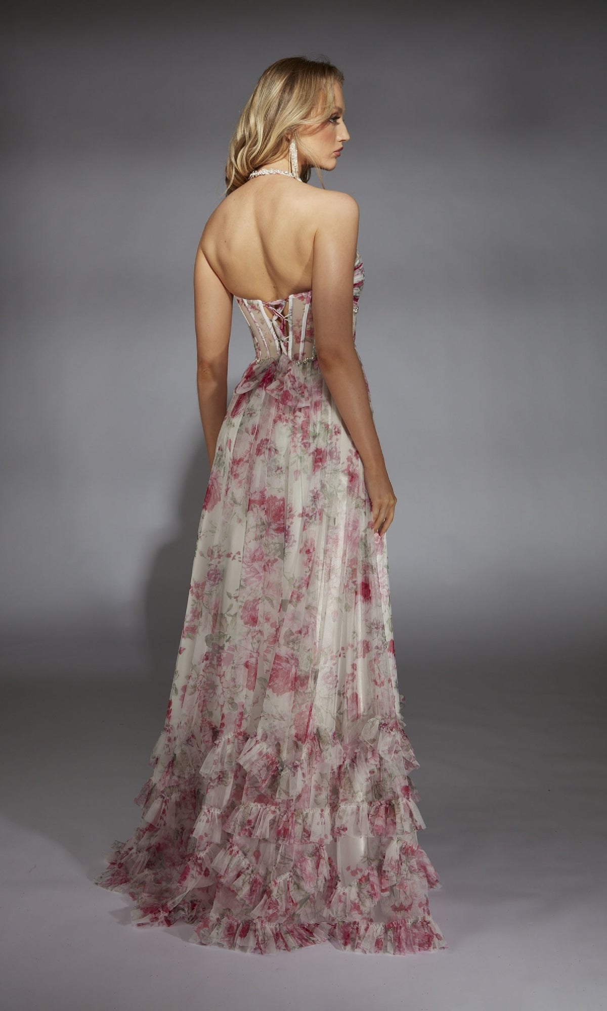 A woman faces away, wearing the Alyce 61757 Long Prom Dress—a strapless, floor-length gown with a floral pattern and ruffled hem—against a plain gray background.
