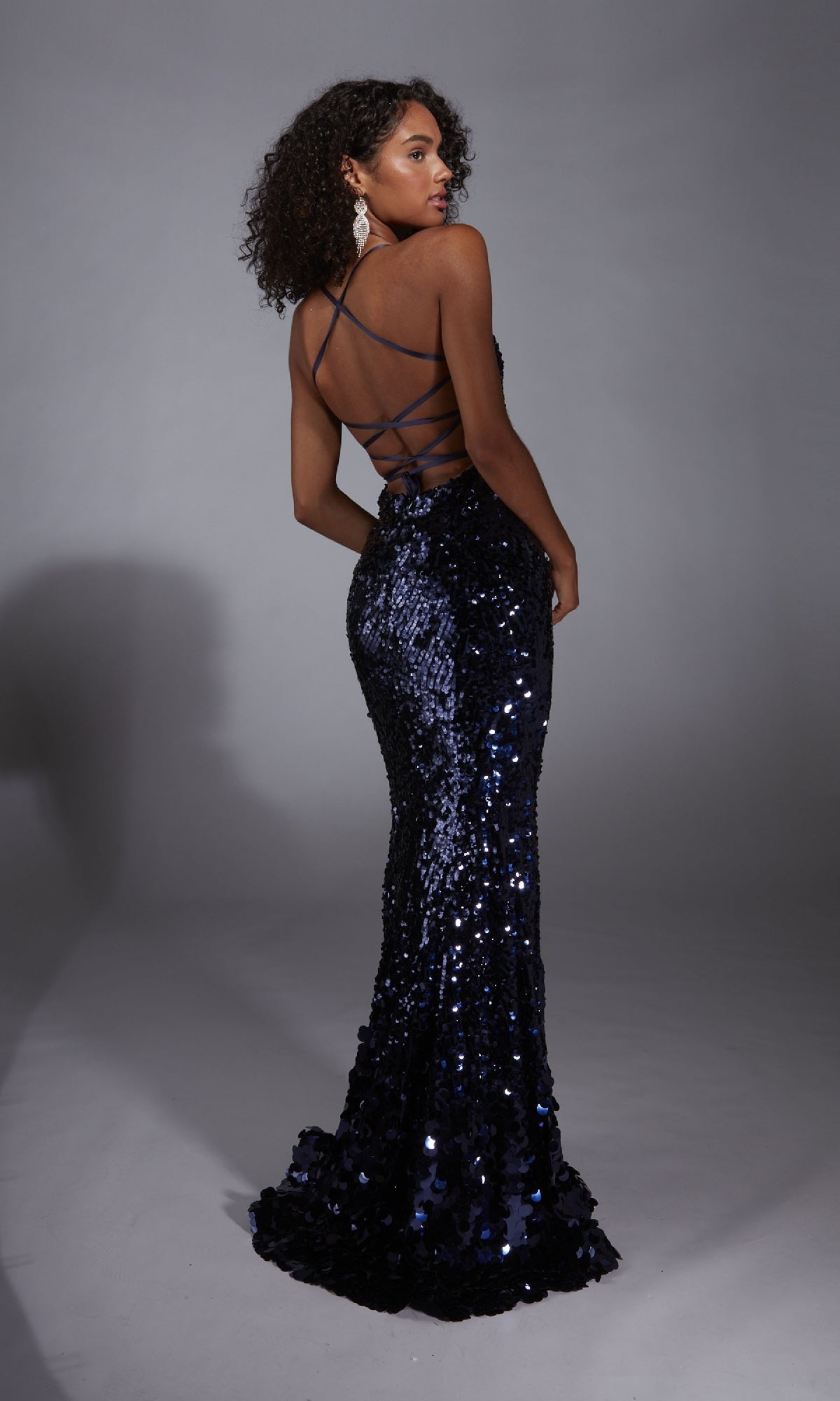 A woman stands against a plain gray background, looking over her shoulder while wearing the Strappy-Back Long Sequin Prom Dress Alyce 61758 in dark blue, featuring an open back with crisscross straps.