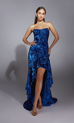 A woman poses in the Alyce 61760, a strapless high-low royal blue sequin prom dress with a train, styled with clear heels and earrings against a plain gray background.