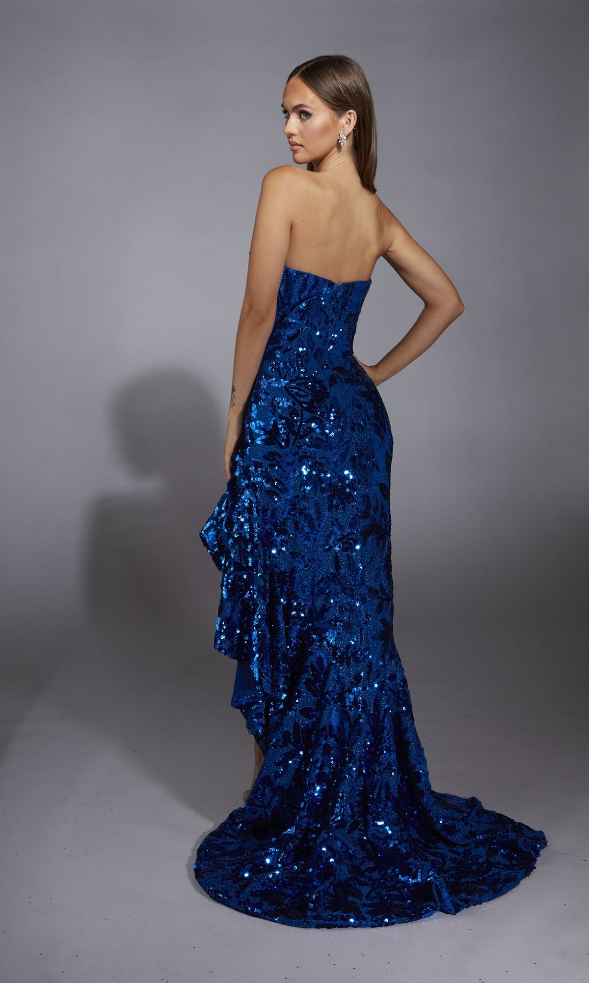 A woman models the Alyce 61760, a royal blue strapless sequin prom dress with a floor-length train, standing before a neutral background and glancing over her shoulder.