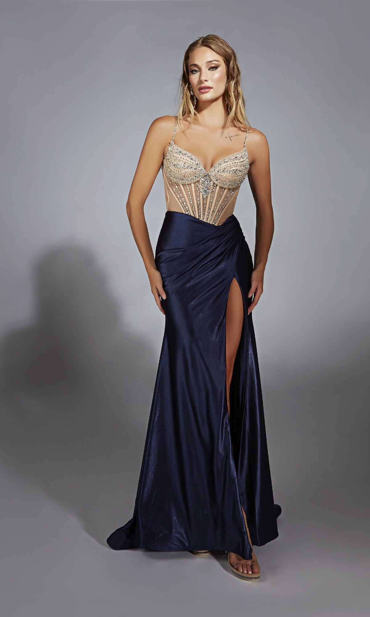 A woman stands in the Long Navy Blue Prom Dress: Alyce 61767, featuring a sleeveless beaded sheer bodice and a dark floor-length skirt with a high leg slit.