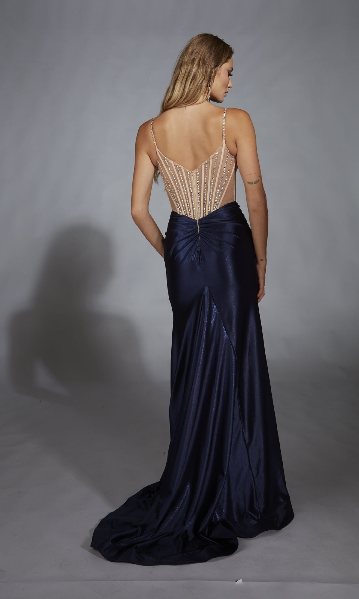 A woman faces away, wearing the Alyce 61767 long navy blue prom dress in stretch satin with a fitted, sheer beaded back and long train, standing against a plain gray backdrop.