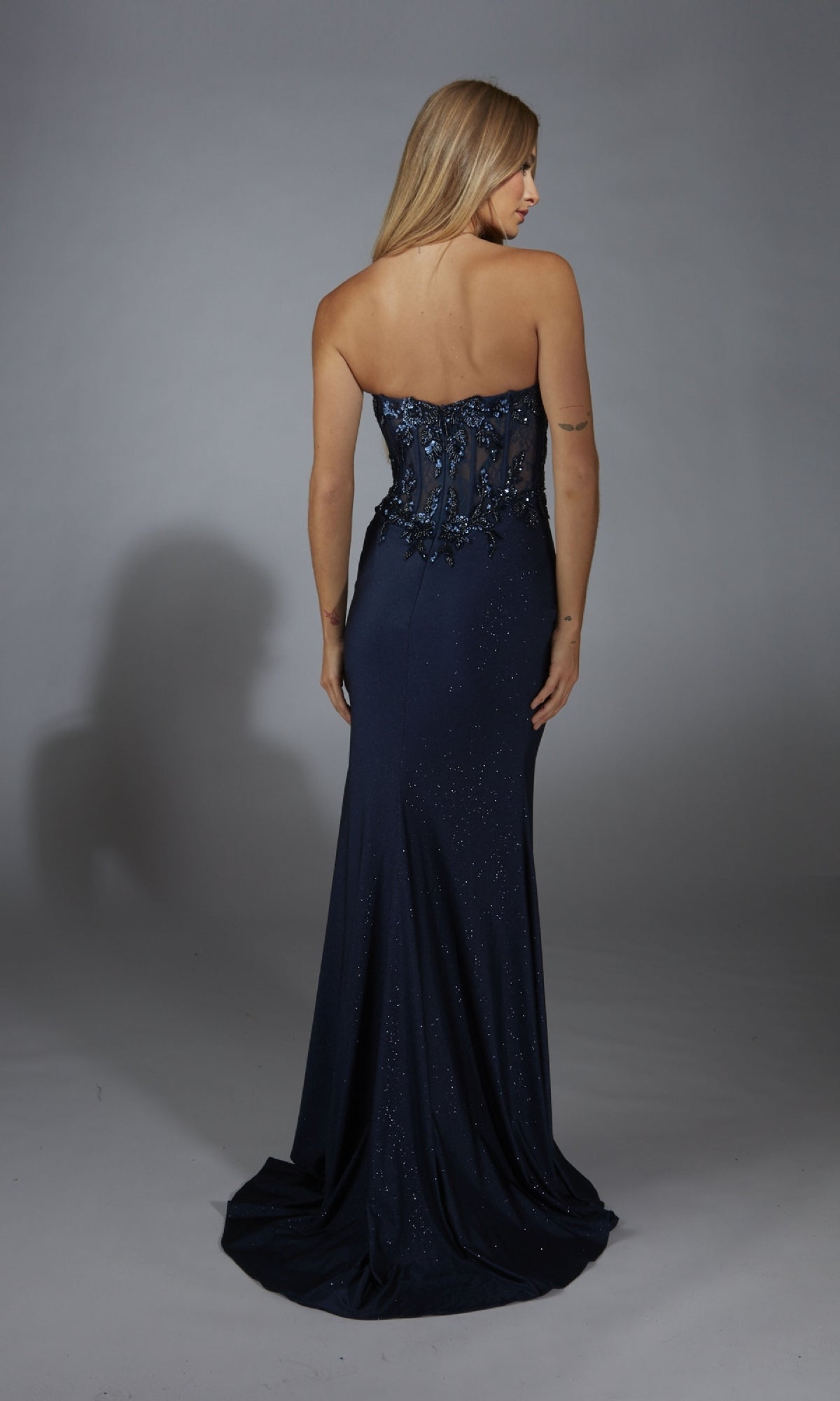 A woman, back turned, models the Alyce 61768 long navy blue prom dress with a strapless sequin bodice and sheer illusion lace corset, set against a plain gray background.