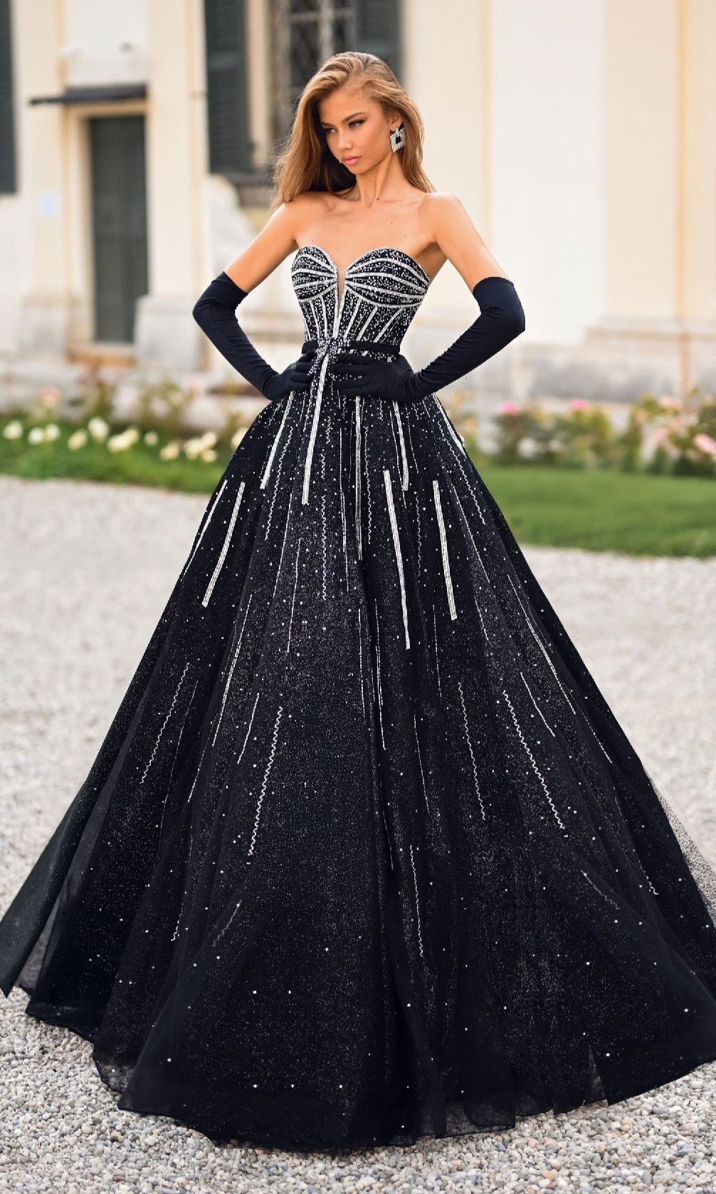 Outdoors, a woman wears the Alyce 61770 long prom dress featuring a strapless glitter corset bodice, a black ball gown skirt with silver embellishments, and elegant black opera gloves.