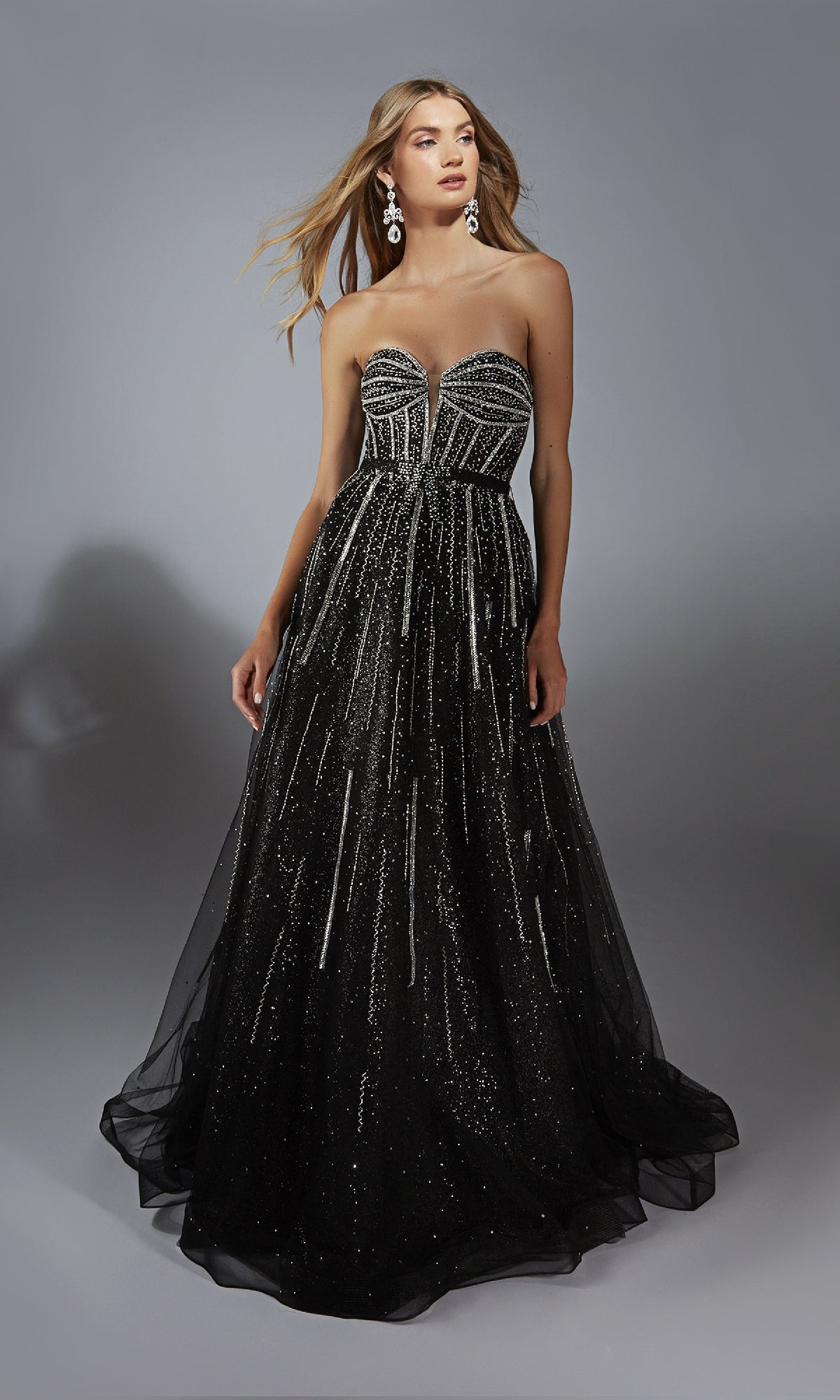 A woman wearing the Alyce 61770 long prom dress, a strapless black gown with a corset bodice, silver beading, and glitter accents stands against a plain gray background.