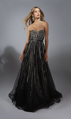 A woman wearing the Alyce 61770 long prom dress, a strapless black gown with a corset bodice, silver beading, and glitter accents stands against a plain gray background.