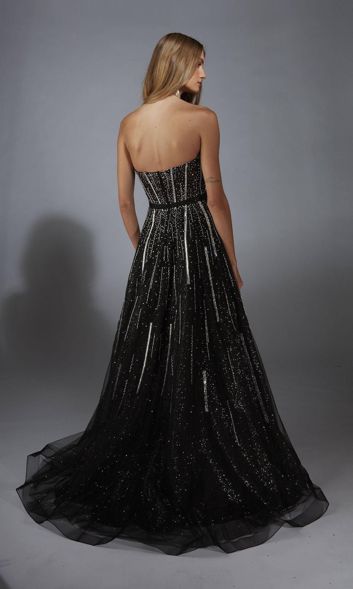 A woman with long blonde hair, facing away from the camera, models the Alyce 61770 long prom dress—a strapless glitter gown with a corset bodice—against a plain gray background.
