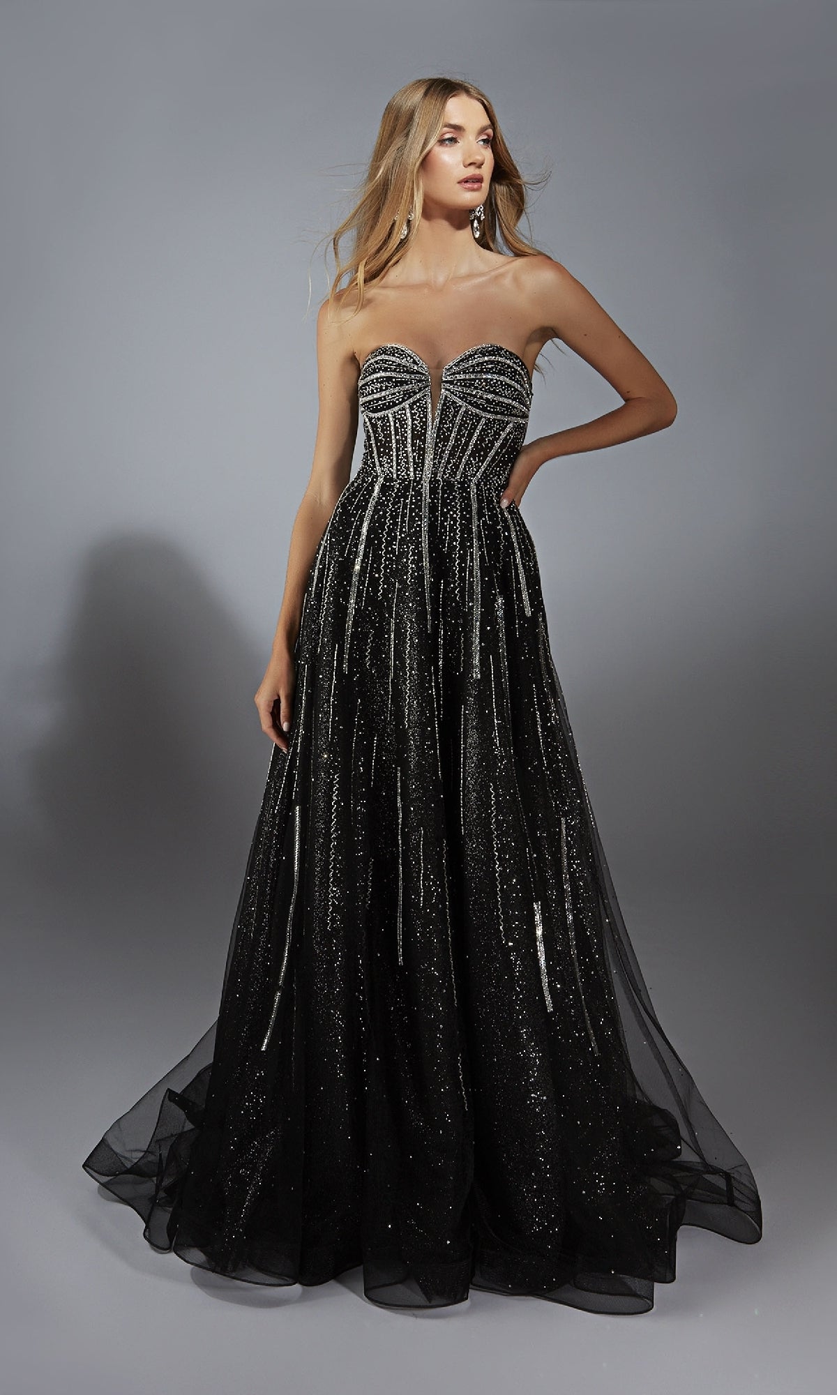 A woman wears the Alyce 61770 long prom dress, featuring a strapless glitter design and corset bodice, standing against a gray background.