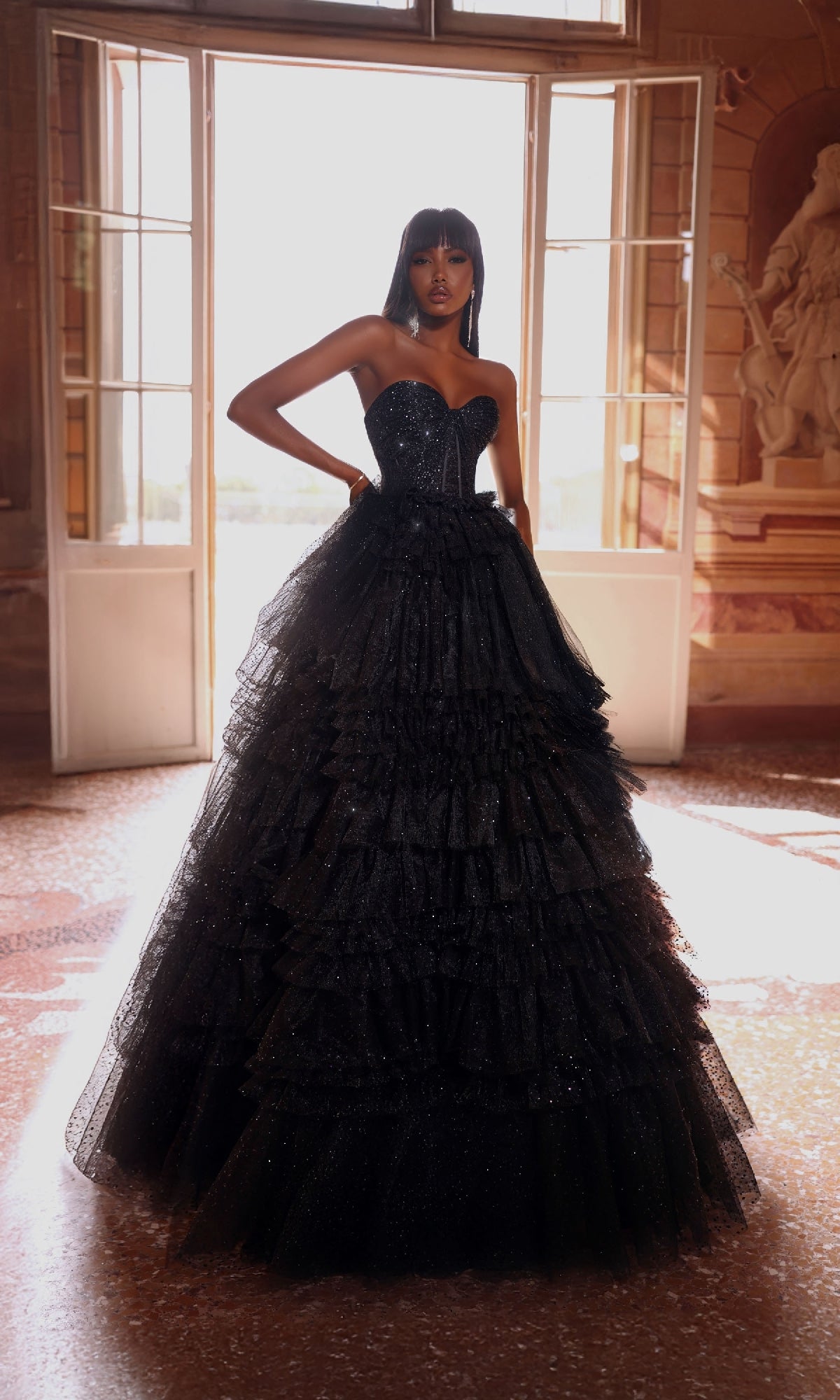 A woman poses indoors in the Alyce 61774 long prom dress, a strapless gown with a fitted bodice and full ruffled skirt, standing before ornate walls and tall open doors for a look of timeless elegance.