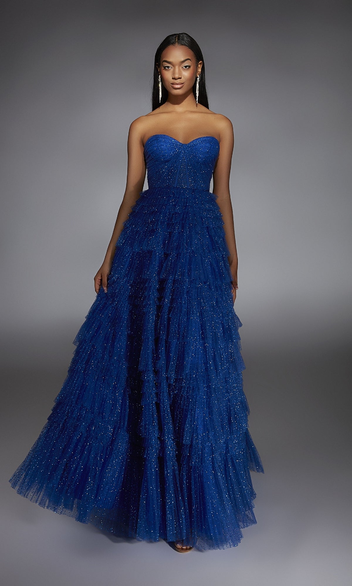 A woman models the Alyce 61774 long prom dress, featuring a strapless corset bodice and layered royal blue fabric with subtle sparkles, standing against a neutral gray background.