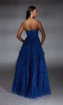 A woman stands with her back turned, wearing the Alyce 61774 long prom dress—strapless, with layered royal blue glitter tulle and sparkling details. She completes the look with long dangling earrings against a neutral background.