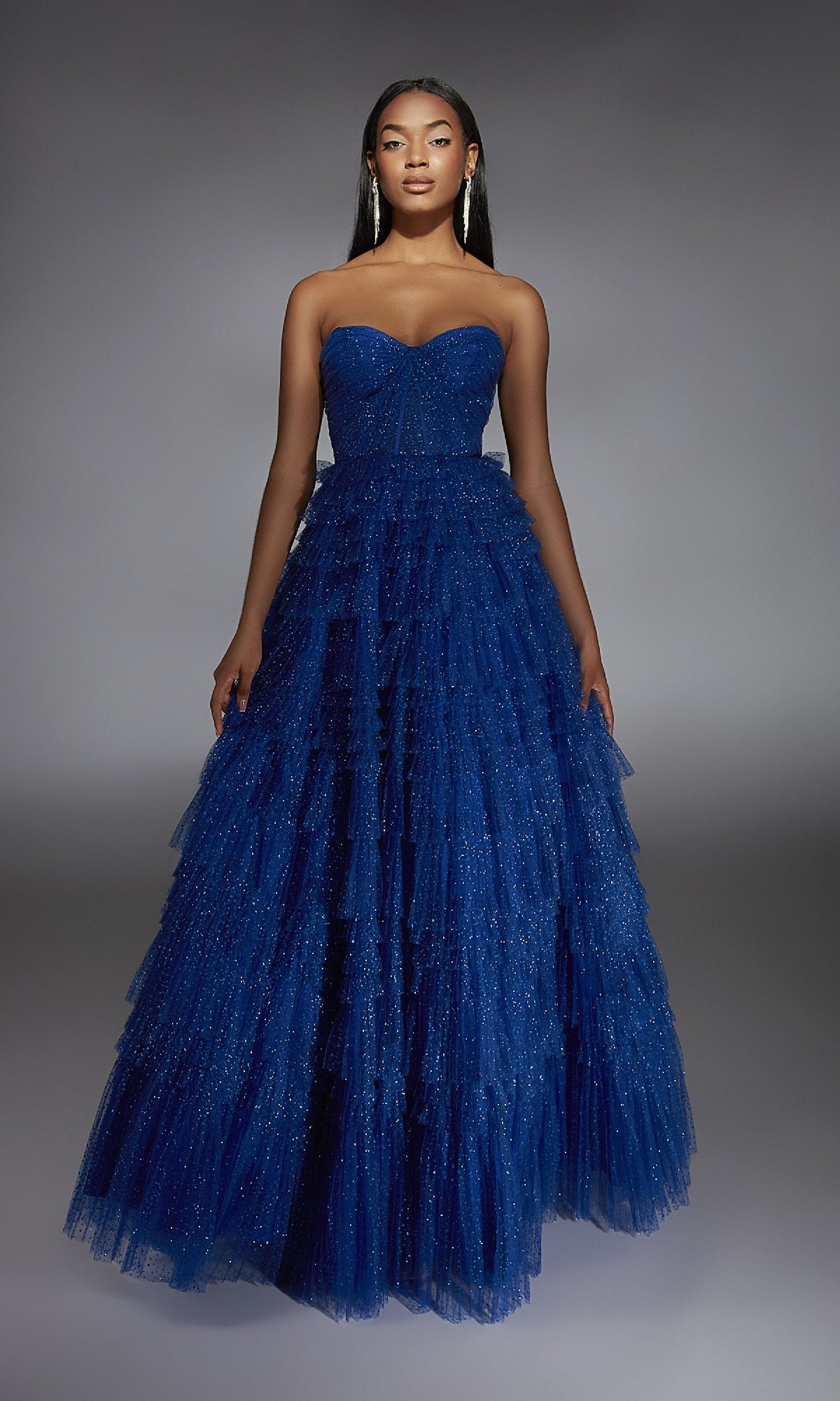 A woman wears the Alyce 61774 long prom dress, a strapless, floor-length deep blue gown with layered tulle and subtle sparkle, standing against a plain grey background.